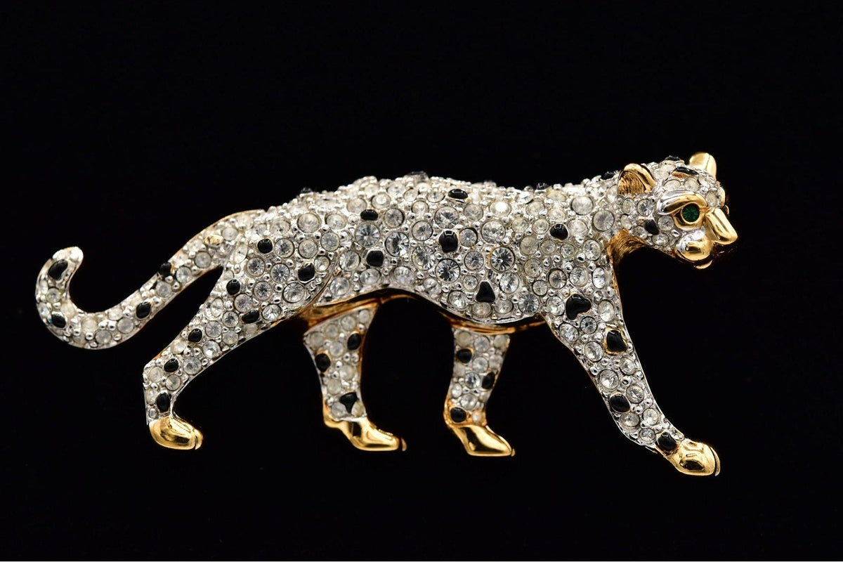 A leopard-shaped brooch or pin covered in clear rhinestones with black spots and gold-tone edges, shown against a solid black background.