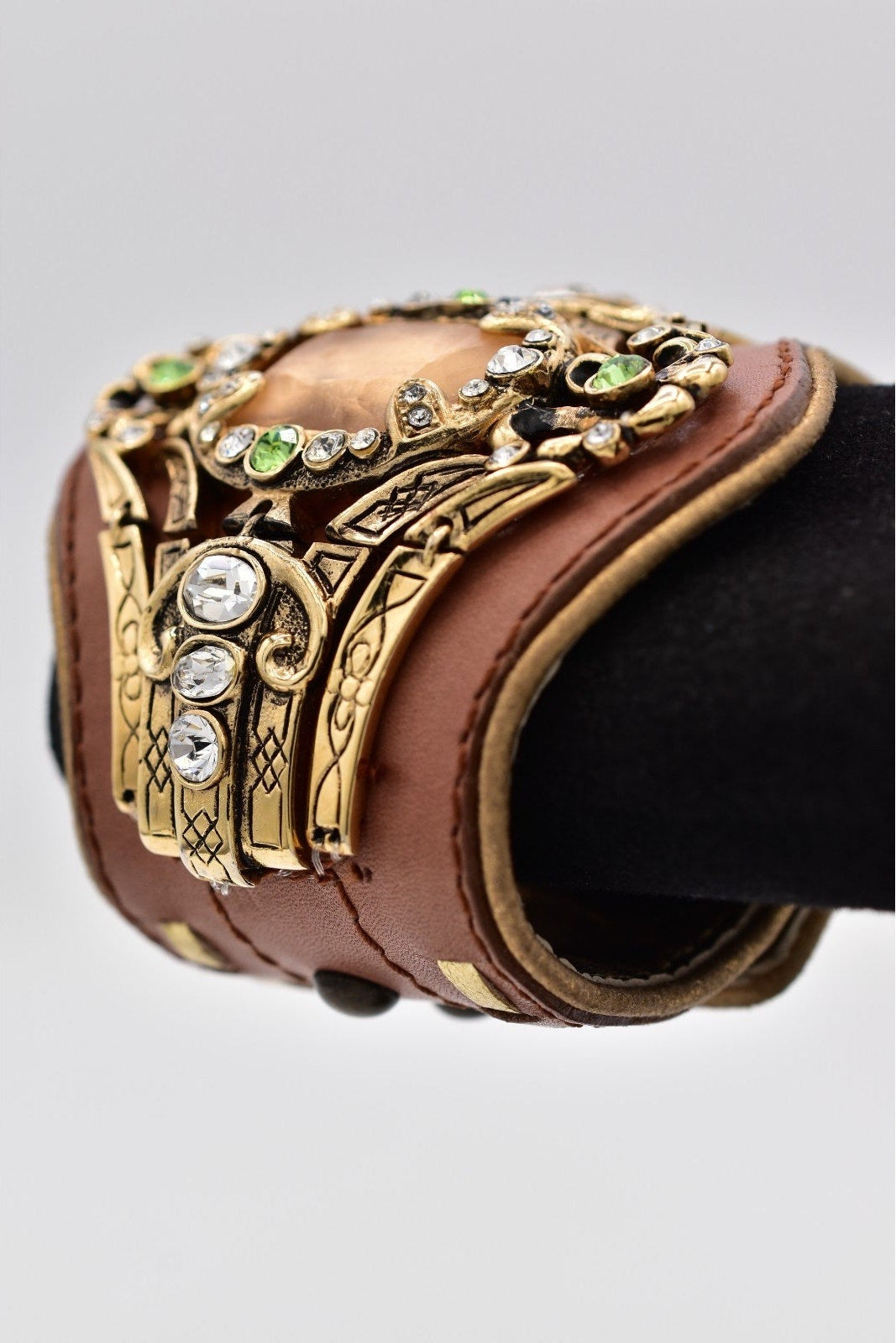 Decorative bracelet with gold accents, gemstones, and brown leather strap on a light gray background