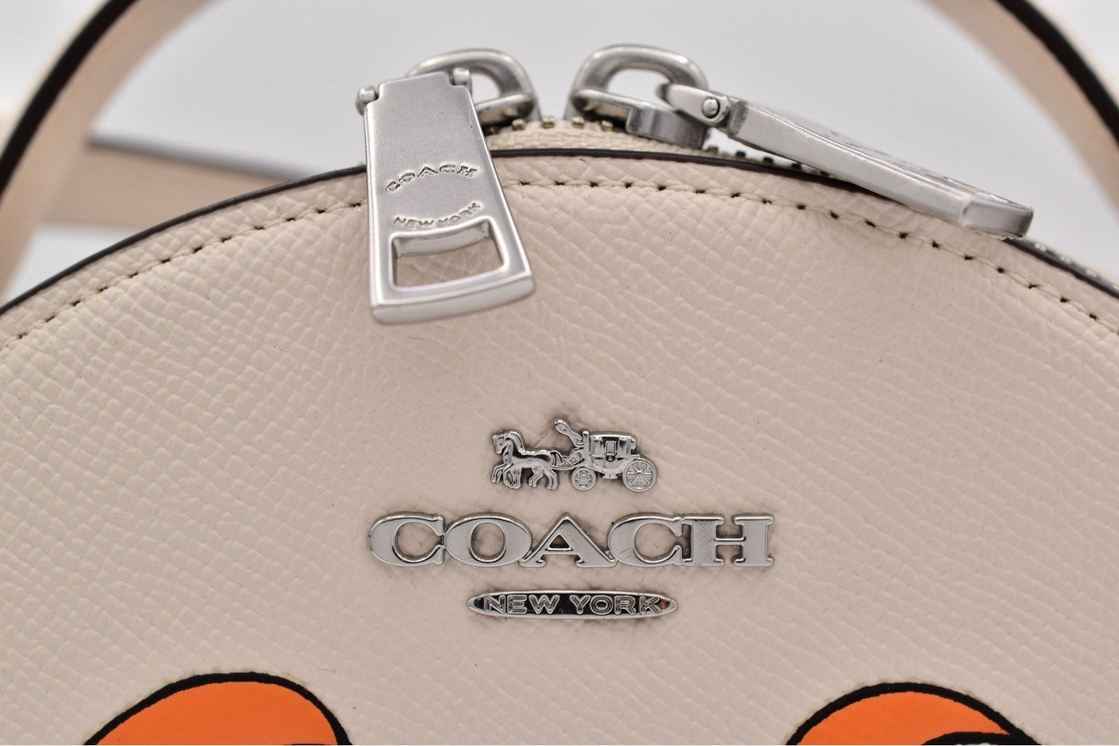 Close-up of a Coach handbag in light beige pebbled leather with a silver-tone zipper pull and the Coach logo on the front.