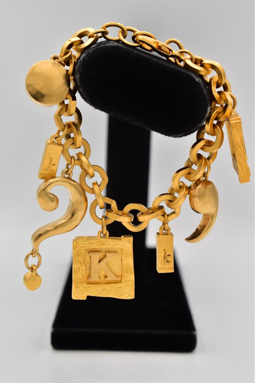 Gold chain bracelet with various charms on a black stand against a gray background