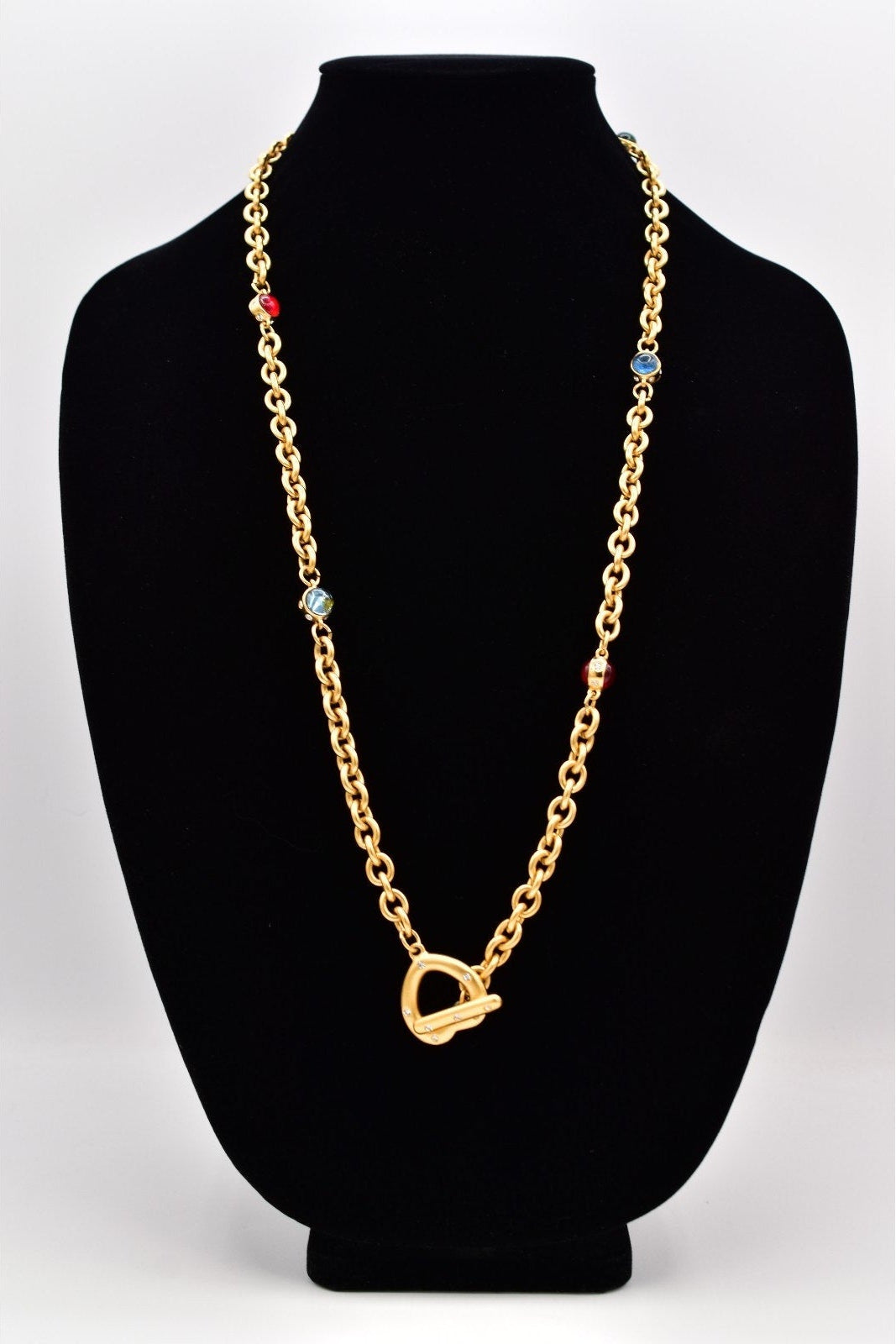 A gold chain necklace with a heart-shaped clasp and small colorful bead accents, displayed on a black jewelry bust against a light gray background.