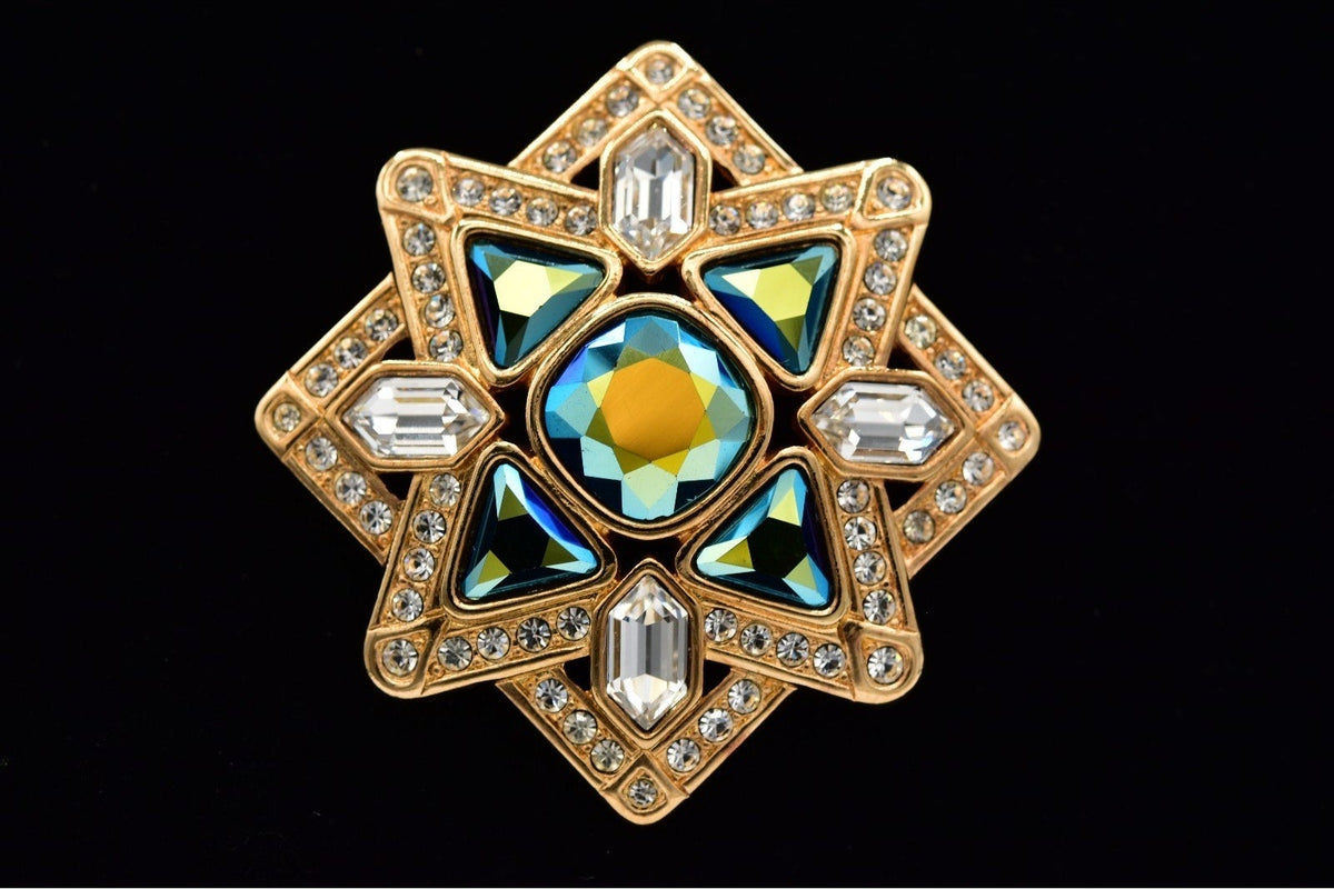 A gold-toned star-shaped brooch featuring a central iridescent blue-green gemstone with a yellow center, surrounded by clear rhinestones and smaller blue-green stones, set against a black background.