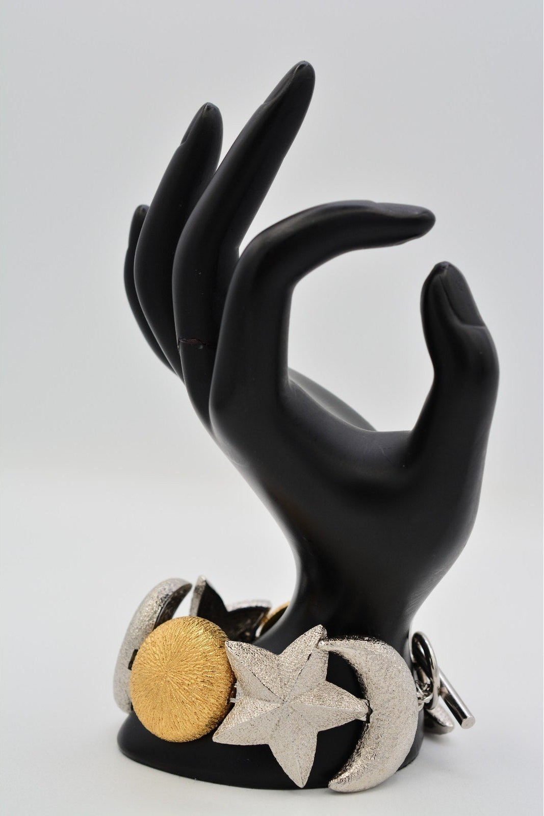 A black hand-shaped jewelry display stand holding a wide bracelet with a textured gold circular element, a silver star-shaped element, and a silver crescent moon element, photographed against a light gray background.