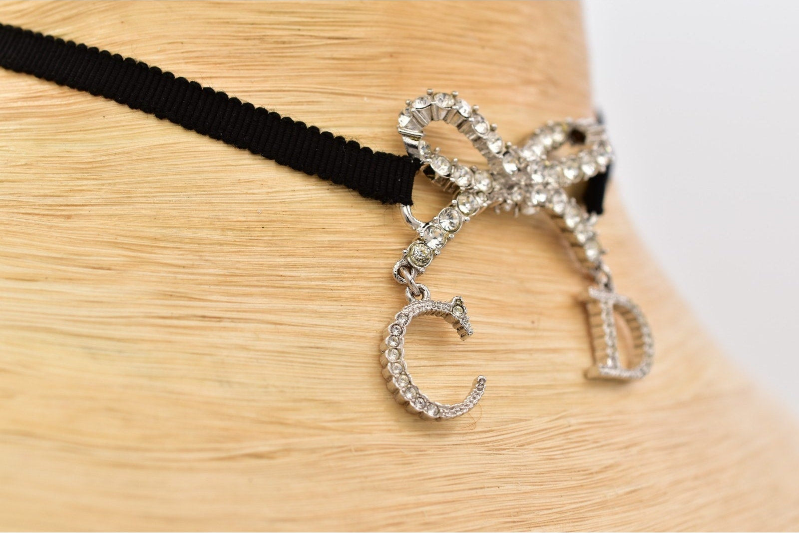 A close-up of a black choker necklace displayed on a light wood mannequin stand. The necklace features a silver-tone bow embellished with clear rhinestones and two dangling rhinestone-encrusted letters, one shaped like a 'C' and the other like a 'D'.