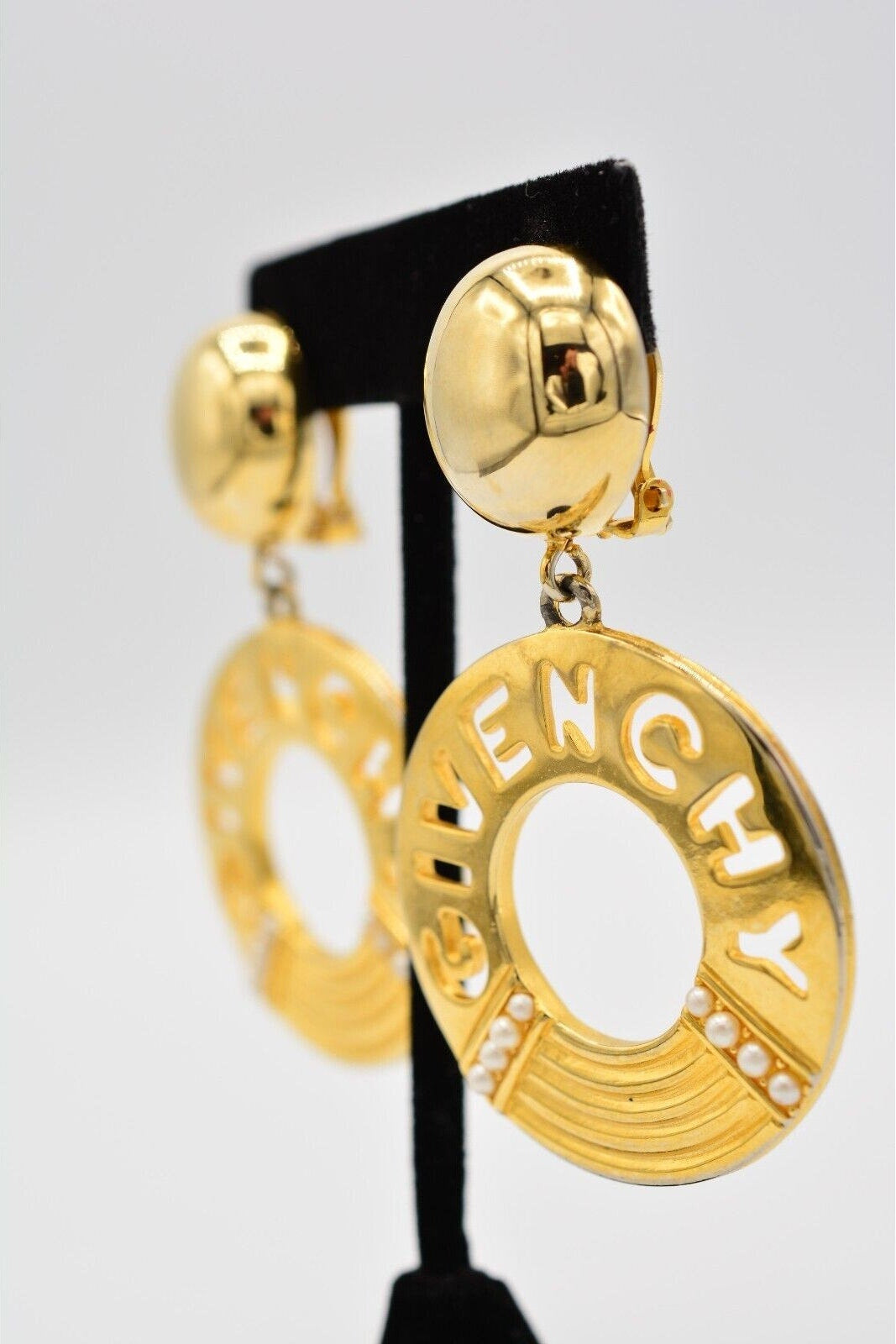 Gold earrings with 'Bvlgari' branding on a black stand against a light gray background