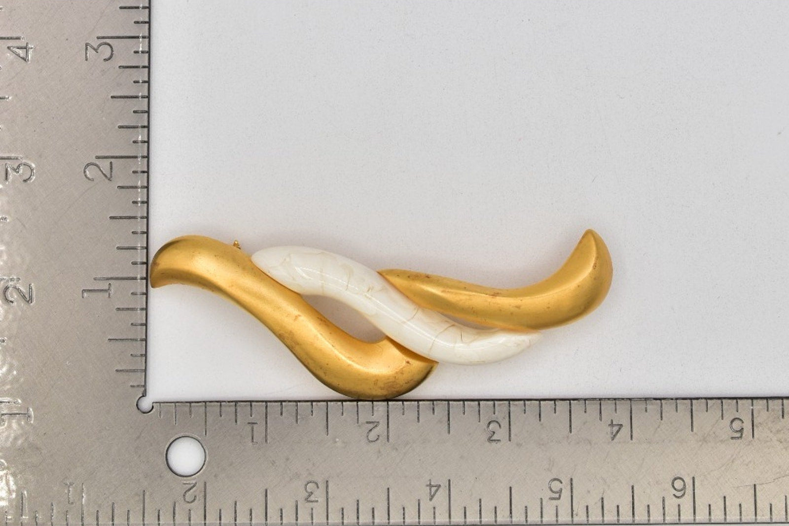 A small decorative object with a gold and white twisted design, shown on a light gray background with a metal ruler at the top left indicating scale.
