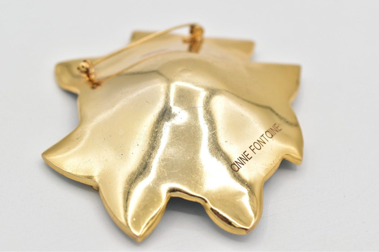 Gold leaf-shaped object with 'ANNE FONTAINE' branding on a light gray background