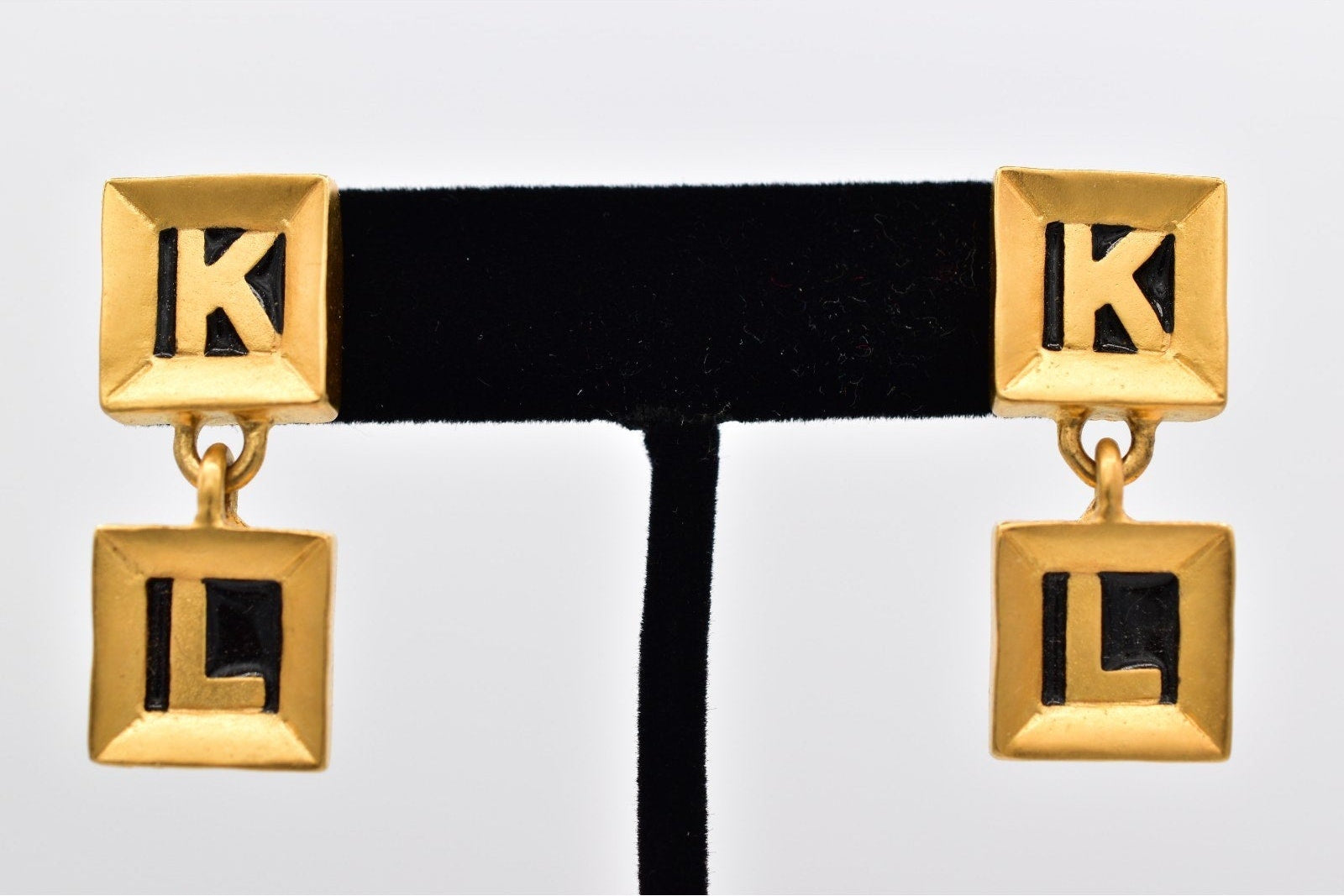 Gold earrings with 'K' and 'L' charms on a black ribbon against a white background