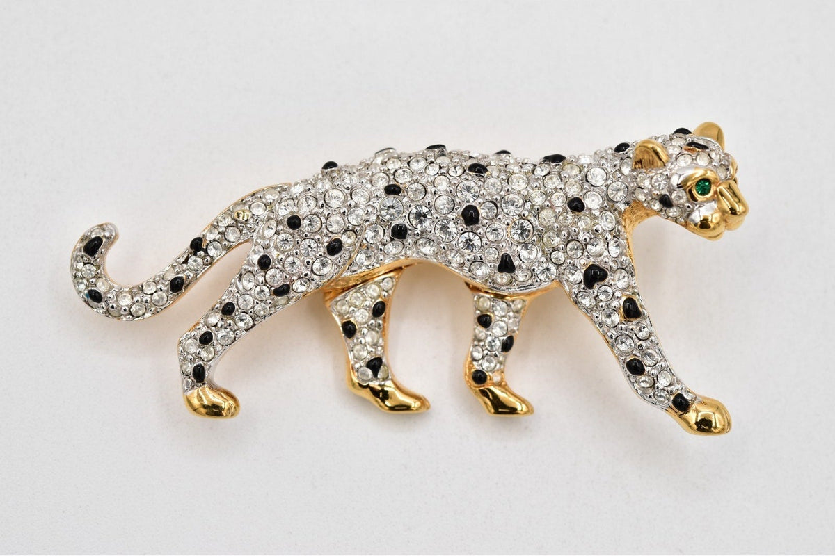 A leopard-shaped brooch or pin featuring a gold-tone body covered in clear rhinestones, with black rhinestones for spots and a small green stone for the eye, shown on a plain light background.